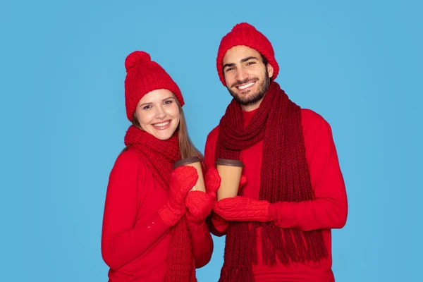 Happy Man And Woman Wearing Mittens Holding Cups With Takeaway Coffee And Smiling At Camera, Joyful Young Couple Drinking Cocoa, Enjoying Celebrating Winter Holidays Together, Blue Background