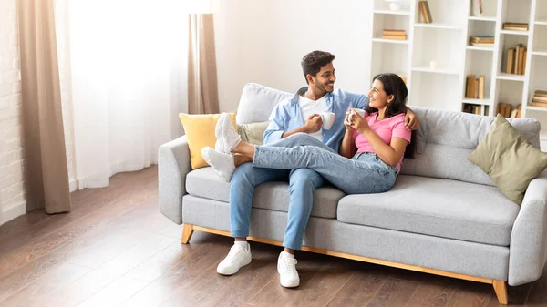 Relaxed indian couple spends a leisurely afternoon on couch, having conversation over cups of coffee. Their homey surroundings envelop them as they share this quiet, intimate moment together.