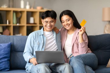 Gülümseyen Asyalı çift evde dizüstü bilgisayarla ödeme yapıyor, bilgisayar ve kredi kartı kullanıyor, evdeki kanepede otururken internet mağazasından alışveriş yapıyor. Modern dijital ticaret
