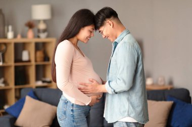Hamilelik Mutluluğu. Genç Koreli bir çiftin bebek beklerken alınlarının birbirine bastırılması, eşinin evde otururken poz verdiği hamile bir kadının karnına dokunması.