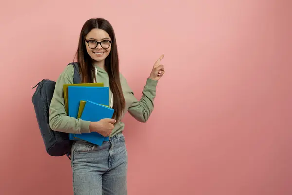看看这个快乐少女手握笔记本 在复印空间边指点 背着书包和眼镜微笑少女 展示粉红背景广告的自由空间 — 图库照片