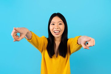 Seni seçiyorum. Sarı tişörtlü, heyecanlı genç Asyalı kadın kamerayı iki eliyle işaret ediyor ve gülümsüyor, mavi stüdyo arka planında izole edilmiş. Koreli kadın.