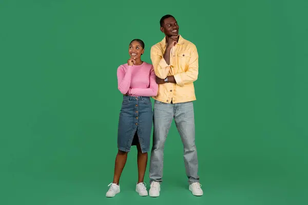 Excited curious young black man and woman wearing casual outfit posing on green studio background, couple standing back to back, touching chin, looking at copy space. Exciting offer, nice proposition
