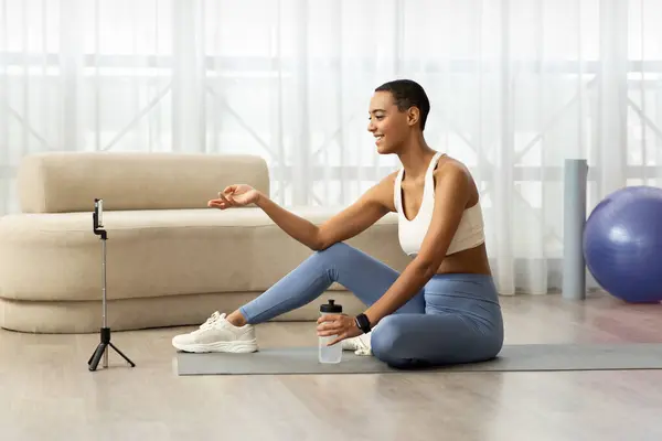 Happy young african american woman blogger in sportswear sit on mat, shooting video fitness blog on phone in living room interior. Health care, weight loss and sports at home, fit lifestyle