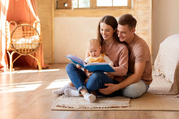 Early Education For Kids. Parents Reading Book To Their Cute Infant Son At Home, Cute Little Baby Looking With Interest, Happy Millennial Family Relaxing Together On Floor In Living Room, Free Space