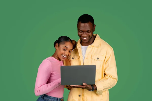 Loving black couple using laptop on green studio background. Happy african american millennial man and woman looking at computer screen and smiling, looking for traveling offers on Internet