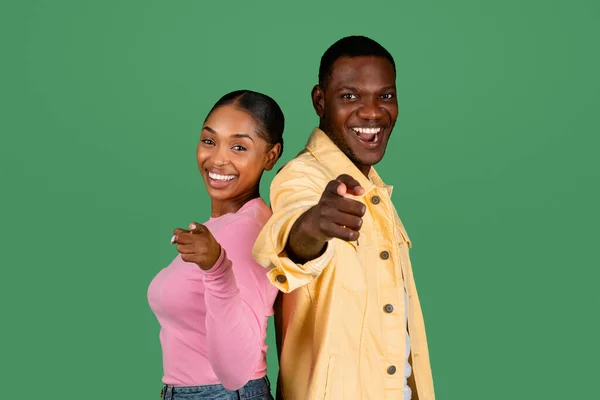 Cheerful positive millennial black couple standing back to back and pointing at camera, isolated on green studio background. Smiling young african american man and woman challenging, inspiring