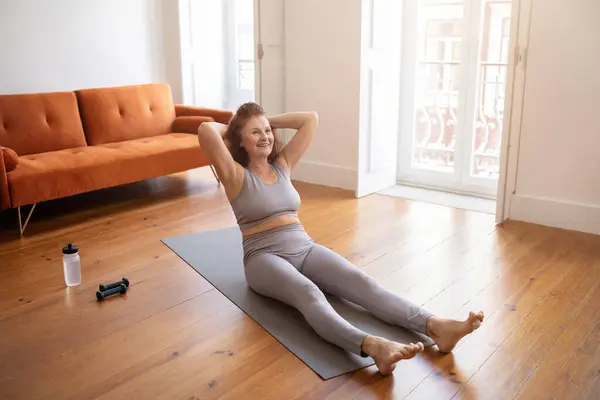 Sporty senior woman training at home, doing abs exercises on yoga mat, smiling active elderly lady working out in living room interior, enjoying domestic sports and healthy lifestyle, copy space