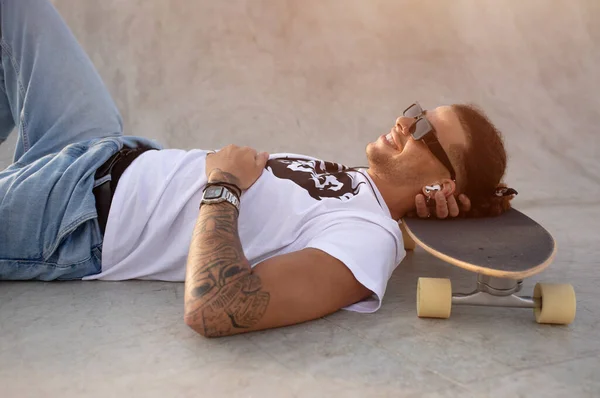 Young european man lying leisurely on his skateboard, outdoors on ramp, finding solace in his music through earpads, side view. The skatepark now serves as a tranquil backdrop for his musical escape.