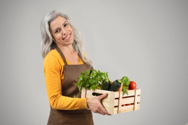Önlüklü neşeli, son sınıf öğrencisi bir kadın. Yanında eko sebzeler olan ahşap bir kutu tutuyor. Gri arka planda izole bir hayat sürüyor. Sağlıklı taze yiyecek, tarım işi, çiftçi işi, reklam ve teklif