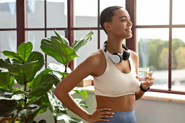 Calm happy young african american woman in sportswear and headphones drink glass of water with lemon, enjoy rest at room interior. Diet at home, healthy lifestyle, peace and weight loss