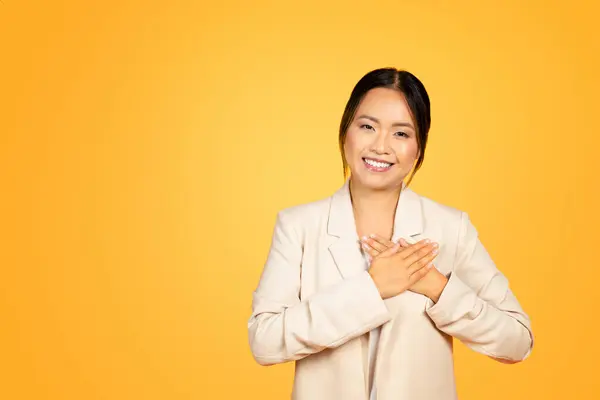 Heartfelt moment of smiling Asian millennial woman places hands on chest, eyes gleaming with genuine gratitude and kindness, isolated on orange studio background. People emotions