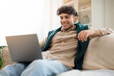 Çevrimiçi Çalışma. Neşeli ortadoğu milenyumu çocuğu elinde kalemle dizüstü bilgisayara bakıyor, internetten eğitim dersleri izliyor, modern oturma odasında bilgisayarla oturuyor.