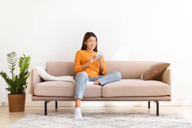 Kanepeye oturmuş, modern cep telefonunu kullanan, flört uygulamasındaki adamlarla sohbet eden, sosyal medyayı kontrol eden, blog okuyan, reklam için kopyalayan güzel Asyalı bir kadın.