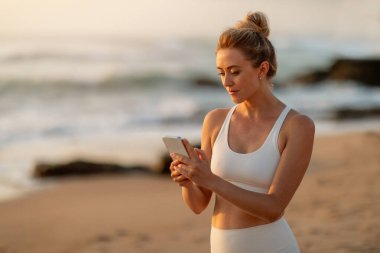 Sahil kenarındaki zinde Avrupalı kadın, gün batımından sonra telefonunu kullanıyor, sakin bir plajda duruyor ve telefon ekranına bakıyor, çevrimiçi antrenörle sohbet ediyor.