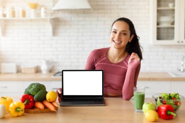 Aktif kıyafetli neşeli bayan ev mutfağında boş beyaz ekran dizüstü bilgisayar gösteriyor. Spor kadını yemek masası yakınındaki bilgisayar ile poz veriyor. Reklam sitesi ya da kilo verme blogu, maket