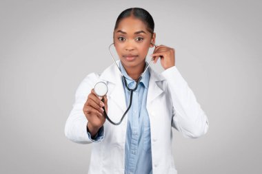 Odaklanmış yetişkin Afrikalı Amerikalı doktor soluğu steteskopla dinler gri arka planda, stüdyoda izole edilir. Tıbbi yardım, sağlık hizmeti, terapistle profesyonel muayene.