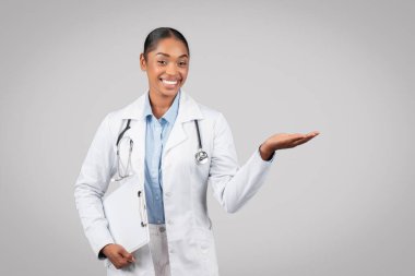 Neşeli yetişkin Afro-Amerikan doktor elinde dosya, gri arka planda, stüdyoda izole edilmiş boş bir yer tutuyor. Tıbbi yardım, sağlık hizmeti tavsiyesi, seçim, reklam ve teklif