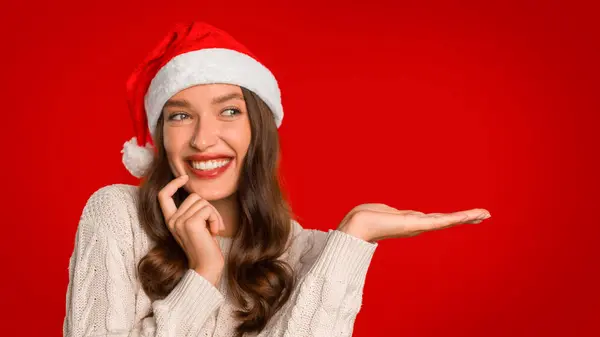 サンタのハットジェスチャーで幸せな若い女性は 赤い背景に対して目に見えないオブジェクトで開いた手を示し クリスマスの休日のオファーや広告製品を提示します パノラマ — ストック写真