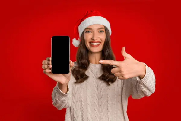 アプリケーション広告のためのモックアップディスプレイで指を指す赤いスタジオの背景にサンタハットに表示スマートフォンの空の画面を示す陽気な若い女性 選択的フォーカス — ストック写真
