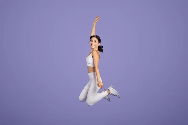 白いアスレチックな衣装のエキサイティングな若い女性は 落ち着いた紫色の背景 完全な長さ 自由なスペースに対して高くジャンプすることによって彼女のエネルギーと活力を示しています — ストック写真