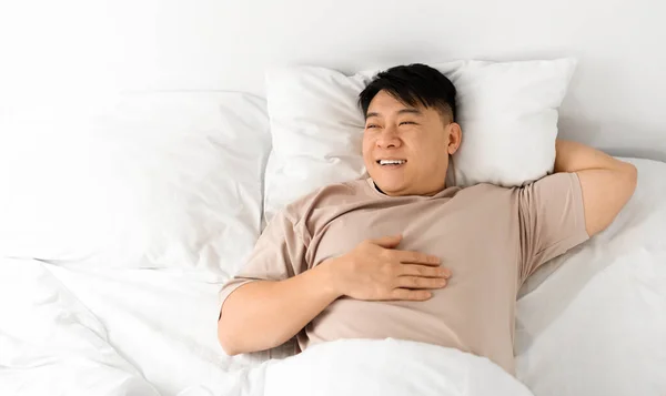 Lazy Morning. Happy Pleased Middle Aged Asian Man Lying In Bed After Nice Sleep, Cheerful Chinese Man Waking Up With Good Mood, Relaxing In Light Bedroom, Enjoying Weekend Pastime, Copy Space