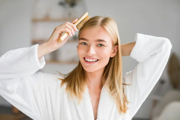 Portrait of happy blonde lady brushing her long hair looking at camera with smile, posing in white bathrobe at bathroom interior. Haircare cosmetics product advertisement