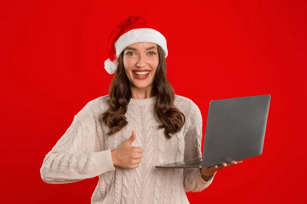 グレートXmasオファー サンタハットで笑顔の女性はラップトップを保持し 赤いスタジオの背景に立って 承認で親指を示しています ウェブサイト — ストック写真