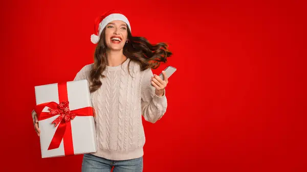 サンタハットの楽しい女性は 赤いスタジオの背景の上に立って携帯電話とクリスマスのプレゼントボックスを保持しています モバイルショッピングアプリケーション オンラインと休日の販売オファー パノラマ 空きスペース — ストック写真