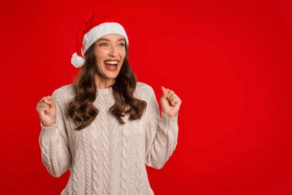 クリスマス休暇 サンタハットで幸せな若い女性は レッドスタジオの背景に立って成功を祝います レディー楽しむXmasのシェイクは喜びと一緒に見て拳を上げました コピースペース — ストック写真