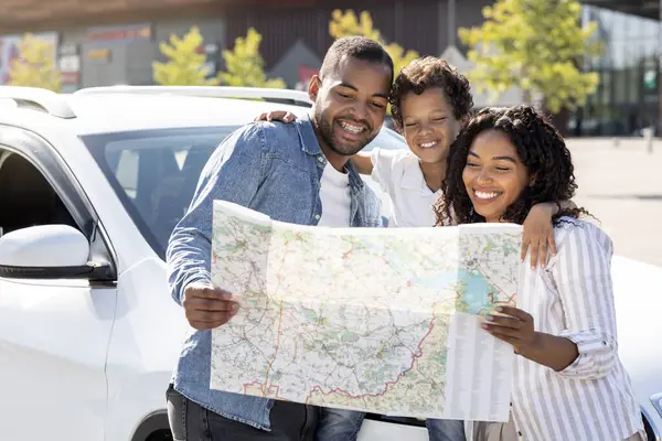 Excited black family of three father mother and preteen son going car trip together at weekend, standing on the street next to white automobile, checking map, choosing for destination to travel