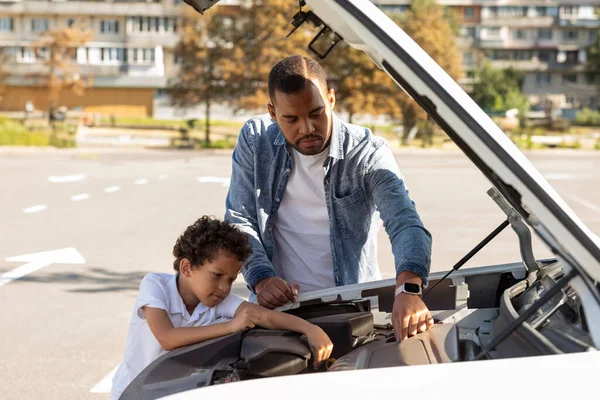 不安的是 黑人爸爸和孩子站在车位上 用破碎的汽车修理引擎 年轻的非洲裔美国父亲教他的小儿子修理家用轿车的一些零件 — 图库照片