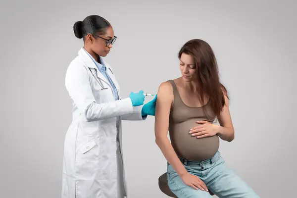 Serious african american lady doctor make injection to pregnant patient during pregnancy, isolated on gray background studio. Vaccination in clinic, medicine, health care, expecting baby