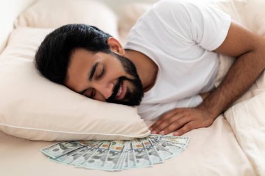 Para Birikimleri. Yastığın altında dolarla uyuyan gülümseyen Hintli adam, rahat bir milenyum erkeği yatakta kestiriyor, banknotlarını evde tutuyor, ekonominin tadını çıkarıyor, yakın plan çekimleri yapıyor.