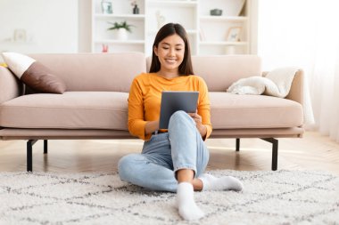Mobil uygulama. Mutlu, genç, Asyalı, günlük kıyafetler giyen bir kadın evdeki oturma odasında oturuyor, dijital tablet kullanıyor ve gülümsüyor, web sörfü yapıyor, arkadaşlarıyla takılıyor, sohbet ediyor, boşluğu kopyalıyor.