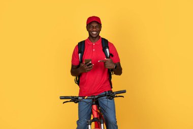 Sırt çantalı, bisikletli, telefonlu, turuncu stüdyo arka planında izole edilmiş neşeli siyah adam kuryesi. Teslimat ve iş için tavsiye uygulaması