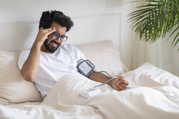 Hypertension Concept. Young Indian Man Checking Blood Pressure With Upper Arm Monitor While Sitting In Bed, Sick Eastern Male Measuring Arterial Tension And Touching Head, Having Acute Headache