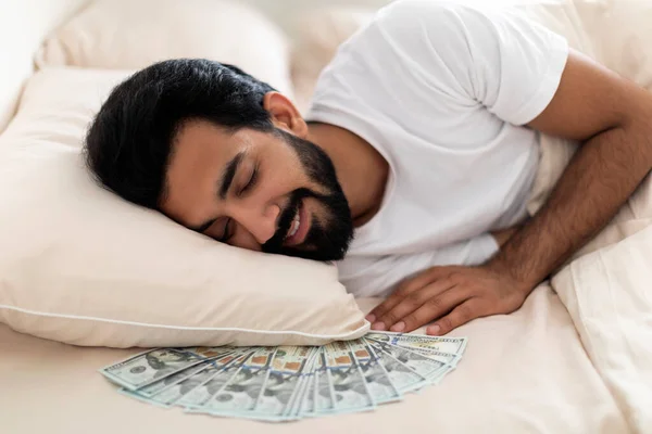 Money Savings. Smiling indian man sleeping with dollar cash under pillow, relaxed millennial eastern male napping in bed, keeping his banknotes at home, enjoying economy, closeup shot