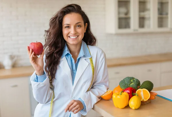 Smiling dietician in white coat presents an apple in front of table filled with colorful fresh fruits and vegetables, promoting a healthy lifestyle