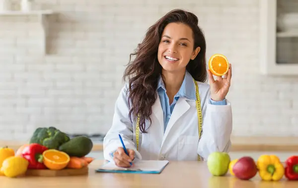 Confident nutritionist in a white coat studies an orange piece, amidst a variety of fresh produce. Shes in the process of drafting a diet chart, with a green smoothie standing nearby