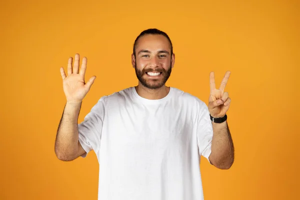 身穿白色T恤的欧洲裔千禧年男子微笑着 一只手拿着五个手指 另一只手拿着和平标志 与橙色背景的工作室相对照 推荐姿势 — 图库照片