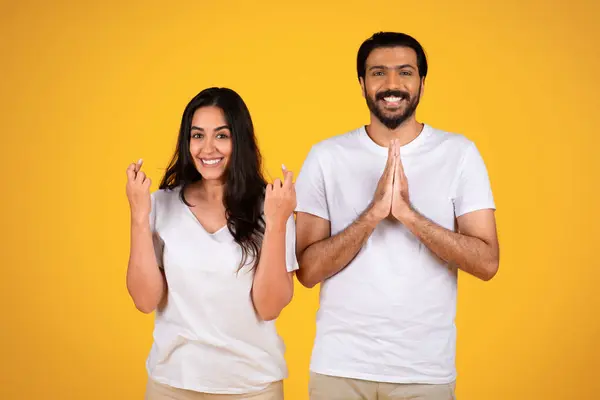 Smiling young arabic couple crossed fingers, make pray gesture, isolated on yellow studio background. Hope, good news, dreams come true and big wish sign to win, people emotions