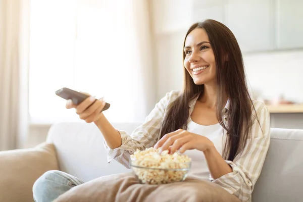 Happy relaxed woman watching tv and eating popcorn at home, switching channels with remote controller, smiling female resting on couch in living room, enjoying domestic weekend, copy space