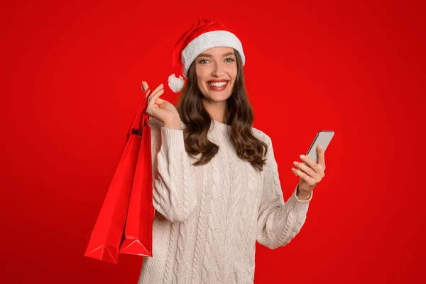 赤い背景に立っているショッピングバッグとスマートフォンを保持しているサンタハットの楽しい女性 電話でXmasの販売をブラウジング幸せバイヤーのスタジオショット モダンな休日のショッピング — ストック写真