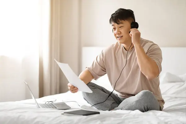 Remote job. Happy handsome middle aged asian man consultant or manager sitting on bed at home, have video call online meeting with team, using computer and headset, reading papers, copy space