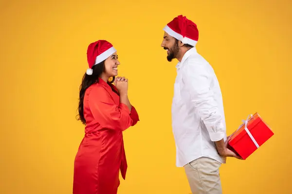 Glad young latin guy in Christmas hat, give red box gift to wife, isolated on yellow background, studio. Joy, present to holiday season, love and celebration New Year together