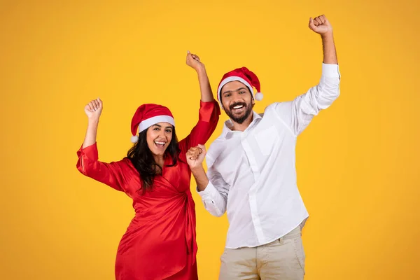 Cheerful latin man and woman in Santa hats, celebrating holiday with raised hands, festive cheer and excitement, dance, isolated on orange studio background. Xmas, New Year mood