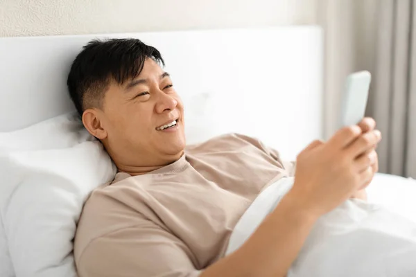 Chinese man using smartphone browsing internet and texting on phone lying in bed at home. Smiling mature asian man in pajamas rest and looks at gadget in white bedroom interior, free space