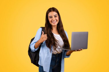 Güzel beyaz bayan öğrencinin dizüstü bilgisayar kullanmasına, el hareketi göstermesine sarı arka planda, stüdyoda izole edilmesine sevindim. Öğrenim onayı, iyi eğitim ve online ders uygulaması, tavsiye işareti