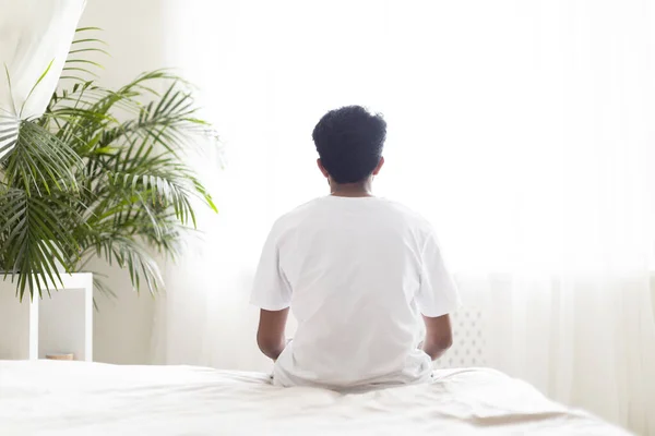 Loneliness Concept. Rear View Of Young Male Sitting On Bed And Looking At Window After Waking Up In The Morning, Unrecognizable Man In White T-Shirt Spending Time In Light Bedroom At Home, Copy Space
